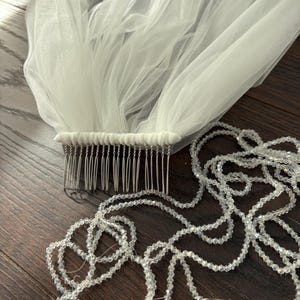 May include: A white tulle bridal veil with a comb attached, and a strand of clear, sparkling beads. The veil is gathered at the comb, creating soft folds. The beads are strung together, forming a delicate, decorative element.