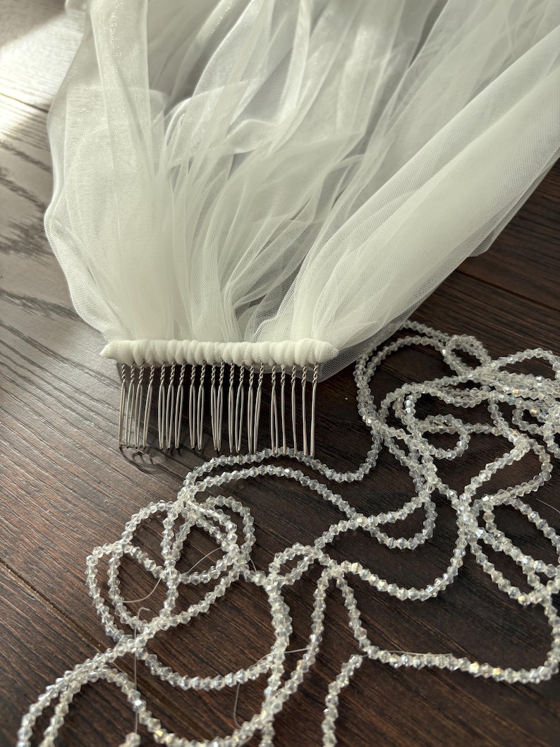 May include: A white tulle bridal veil with a comb attached, and a strand of clear, sparkling beads. The veil is gathered at the comb, creating soft folds. The beads are strung together, forming a delicate, decorative element.