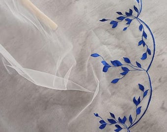 May include: White sheer fabric with a blue embroidered floral design. The fabric has a scalloped edge.
