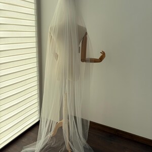 May include: A long, sheer white bridal veil is draped over a mannequin. The veil cascades down the back, creating a flowing effect. The mannequin is positioned near a window with blinds, and the floor is dark wood.