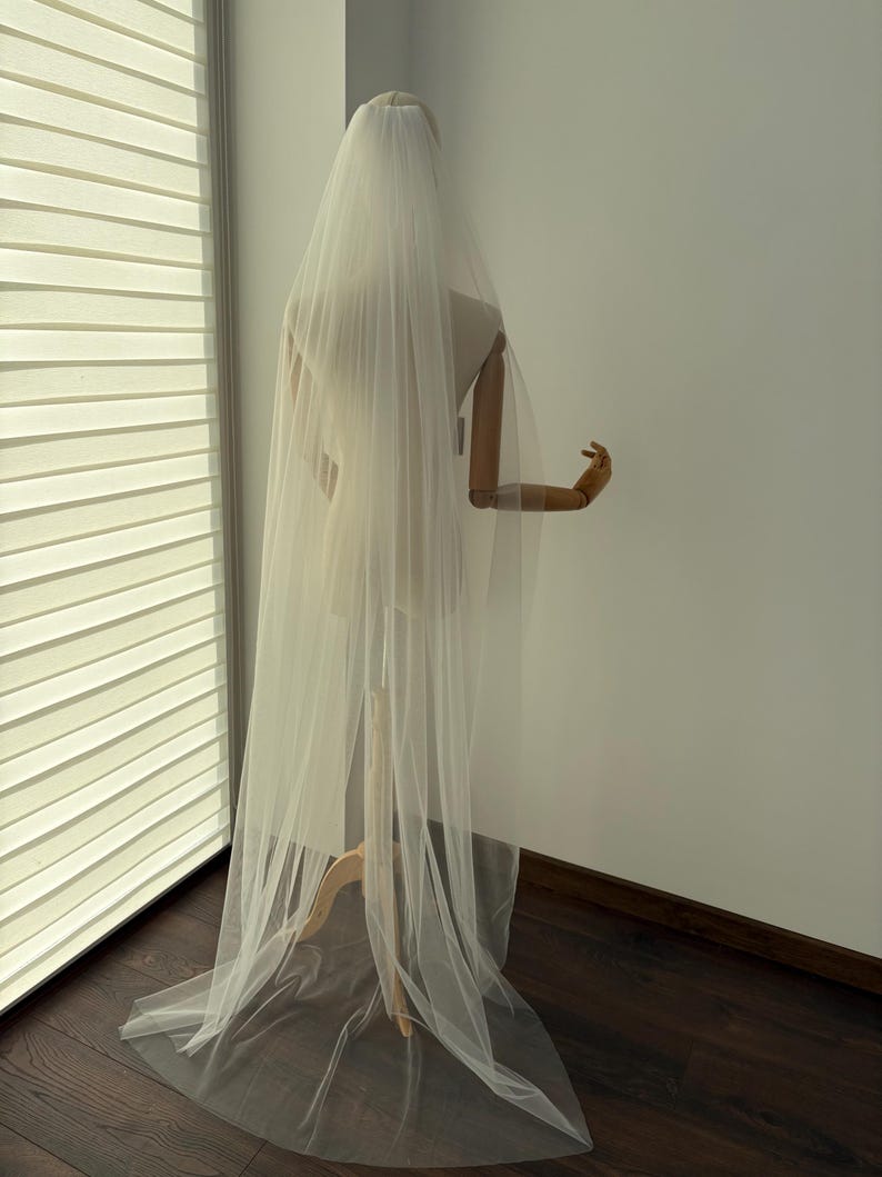 May include: A long, sheer white bridal veil is draped over a mannequin. The veil cascades down the back, creating a flowing effect. The mannequin is positioned near a window with blinds, and the floor is dark wood.