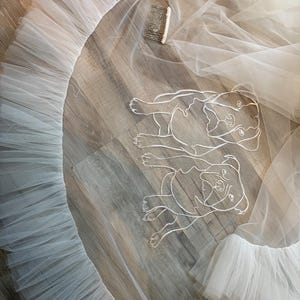 May include: A white tulle wedding veil with a ruffled edge and a comb. The veil features an embroidered outline of two dogs. The veil is lying on a wooden surface.