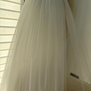 May include: A sheer, ivory-colored bridal veil. The veil is made of a lightweight, flowing fabric, and is attached to a headpiece. The veil is draped over a mannequin head, and the fabric is slightly translucent, allowing light to pass through.