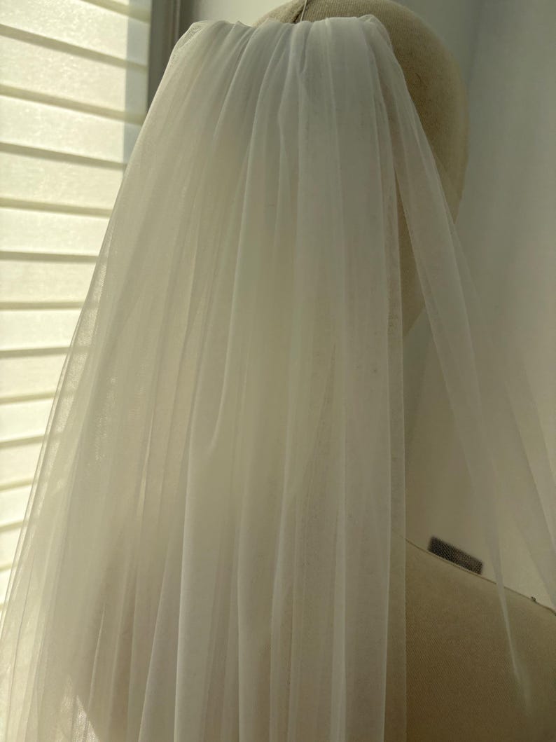 May include: A sheer, ivory-colored bridal veil. The veil is made of a lightweight, flowing fabric, and is attached to a headpiece. The veil is draped over a mannequin head, and the fabric is slightly translucent, allowing light to pass through.