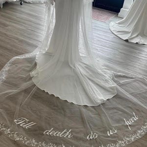 May include: A white wedding dress with a long train and a sheer veil. The veil is embroidered with the words "Till death do us part" and a floral design along the edge. The dress is in a bridal shop.