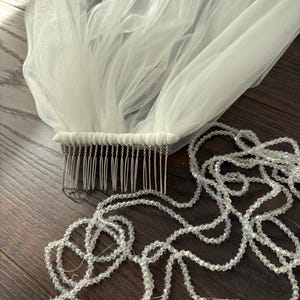May include: A white bridal veil with a comb and a strand of clear, faceted beads. The veil is made of a sheer, flowing fabric. The comb is silver-toned and attached to the veil. The beads are strung together in a long, delicate strand.