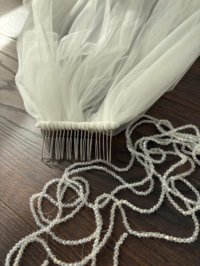 May include: A white bridal veil with a comb and a strand of clear, faceted beads. The veil is made of a sheer, flowing fabric. The comb is silver-toned and attached to the veil. The beads are strung together in a long, delicate strand.