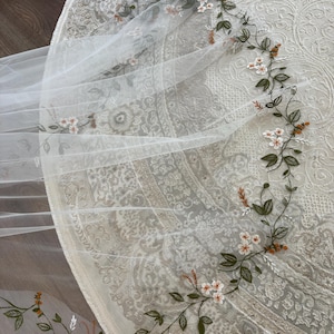 Può includere: Un velo in tulle bianco trasparente con un delicato ricamo di viticci verdi, piccoli fiori bianchi con centri arancioni e minuscoli boccioli arancioni. L'intricato motivo floreale segue il bordo curvo del tessuto in un disegno continuo.