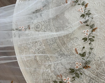May include: A sheer white tulle veil featuring delicate embroidery of winding green vines, small white flowers with orange centers, and tiny orange buds. The intricate floral pattern follows the curved edge of the fabric in a continuous design.