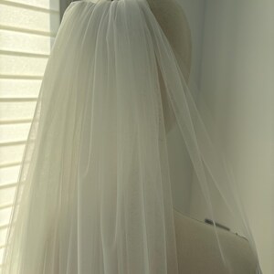 May include: A sheer, white bridal veil is draped over a mannequin head. The veil is made of a delicate, flowing fabric, and is attached to the head with a pin. The veil cascades down, creating a soft, ethereal look.