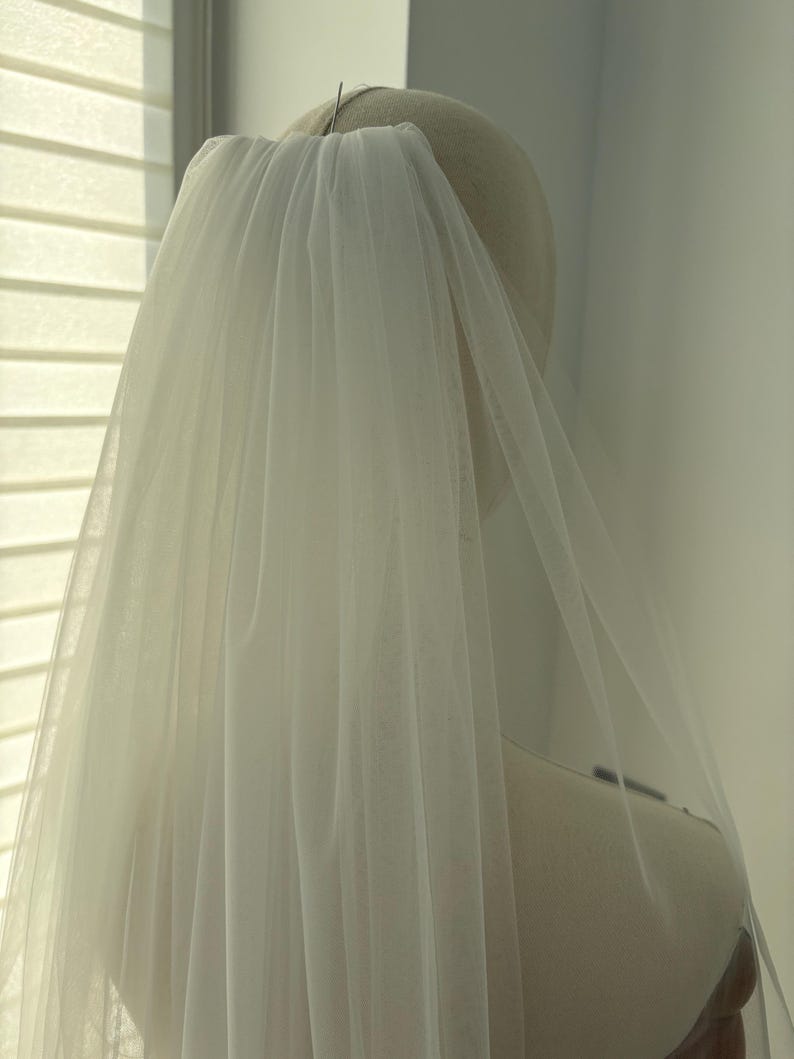 May include: A sheer, white bridal veil is draped over a mannequin head. The veil is made of a delicate, flowing fabric, and is attached to the head with a pin. The veil cascades down, creating a soft, ethereal look.