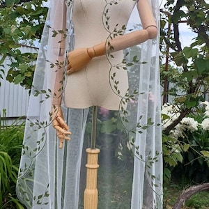 May include: White bridal veil with a delicate green leaf pattern embroidered around the edges. The veil is displayed on a wooden stand.