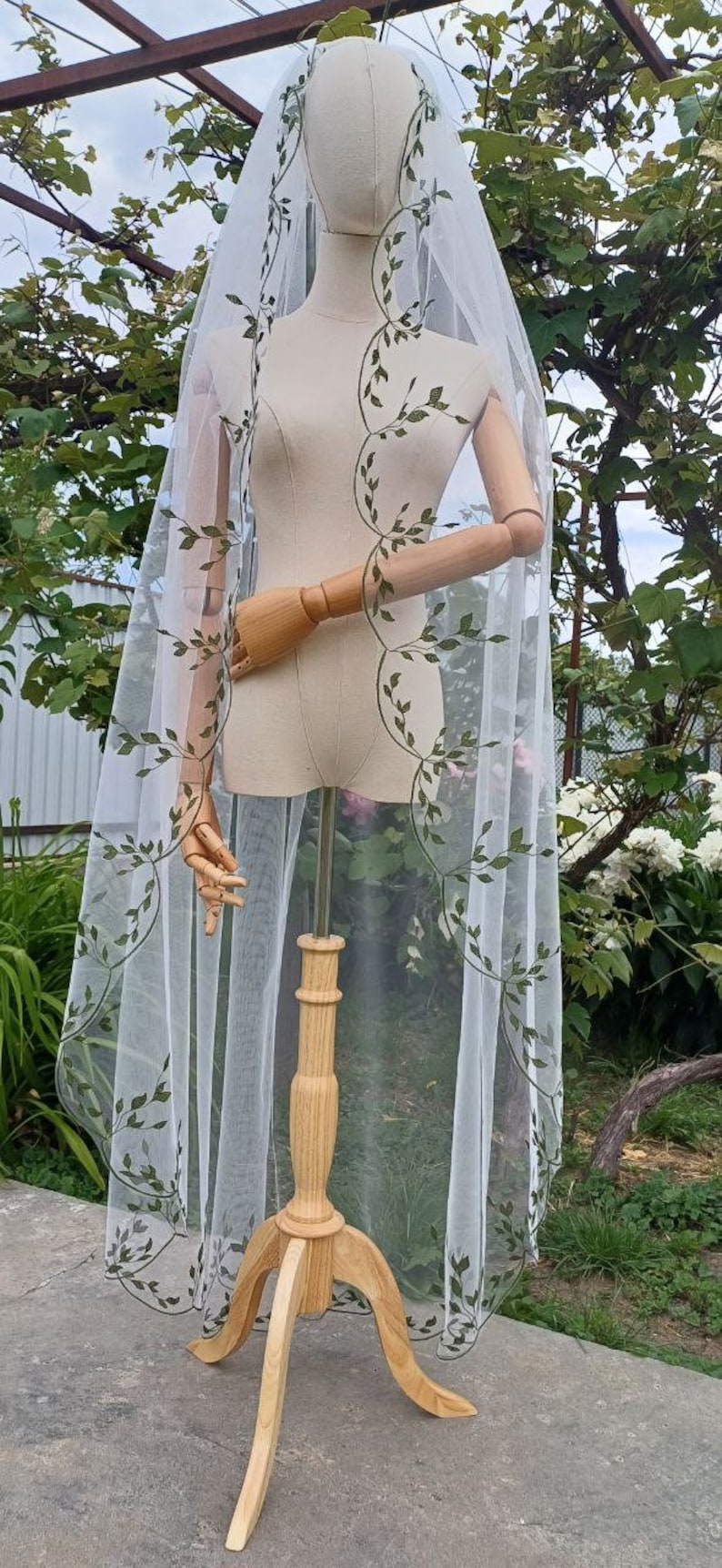 May include: White bridal veil with a delicate green leaf pattern embroidered around the edges. The veil is displayed on a wooden stand.