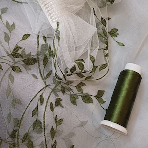 May include: White tulle fabric with green embroidered leaves. A spool of green thread is on the fabric.