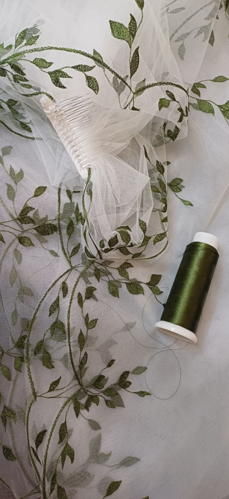 May include: White tulle fabric with green embroidered leaves. A spool of green thread is on the fabric.