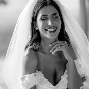 May include: A black and white photograph of a bride wearing a white off-the-shoulder wedding dress with floral accents. She is smiling with her eyes closed, and a sheer veil drapes over her head. A ring is visible on her finger.