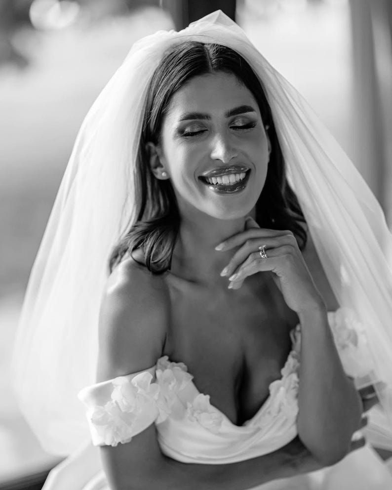 May include: A black and white photograph of a bride wearing a white off-the-shoulder wedding dress with floral accents. She is smiling with her eyes closed, and a sheer veil drapes over her head. A ring is visible on her finger.