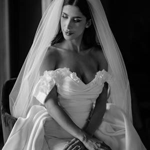 May include: A black and white photograph of a bride wearing a white, off-the-shoulder wedding dress with floral accents. A sheer veil drapes over her dark hair. She is seated, with her hands clasped in her lap, and a ring is visible on her finger.