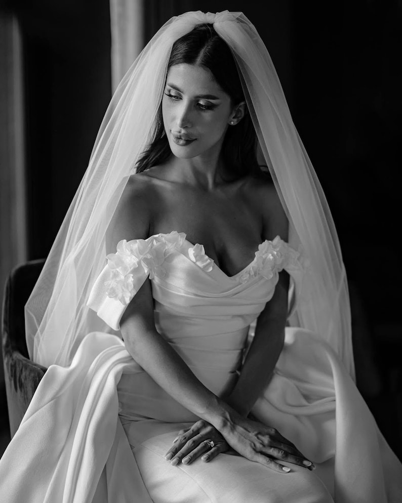 May include: A black and white photograph of a bride wearing a white, off-the-shoulder wedding dress with floral accents. A sheer veil drapes over her dark hair. She is seated, with her hands clasped in her lap, and a ring is visible on her finger.