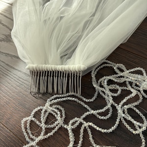 May include: A white tulle bridal veil with a comb attached, and a strand of clear, faceted beads. The veil is gathered at the comb, creating a flowing, layered effect. The beads are strung together, adding a touch of sparkle.