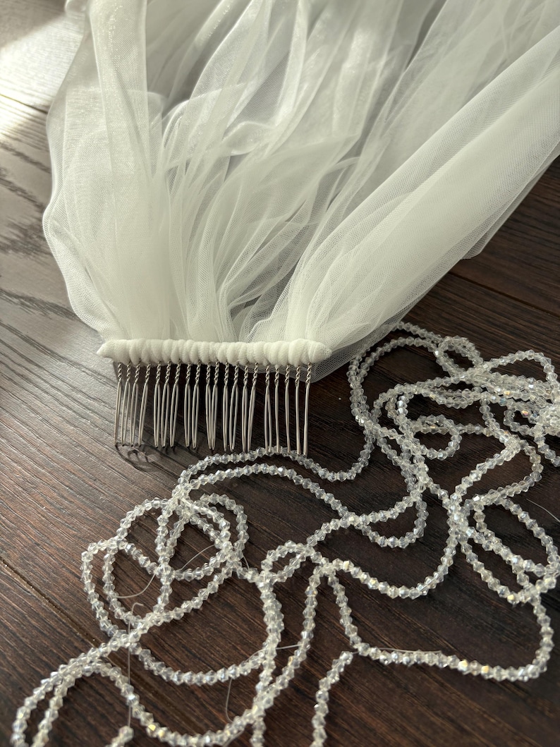 May include: A white tulle bridal veil with a comb attached, and a strand of clear, faceted beads. The veil is gathered at the comb, creating a flowing, layered effect. The beads are strung together, adding a touch of sparkle.