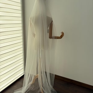 May include: A long, sheer white bridal veil draped over a mannequin. The veil cascades down, creating soft folds and a flowing effect. The mannequin is positioned near a window with blinds, and the background is a plain white wall.