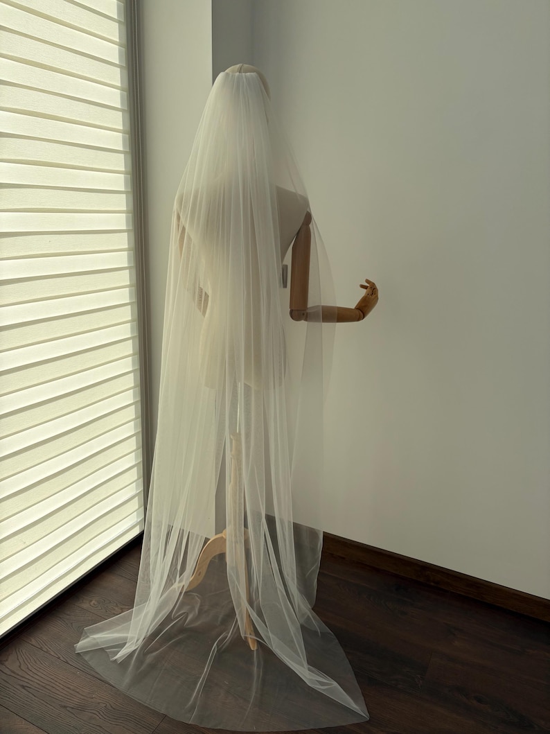 May include: A long, sheer white bridal veil draped over a mannequin. The veil cascades down, creating soft folds and a flowing effect. The mannequin is positioned near a window with blinds, and the background is a plain white wall.