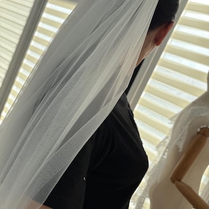 May include: A sheer white bridal veil, draped over the head and shoulders. The veil is made of a delicate, flowing fabric, creating a soft, ethereal look. The image is taken from a side angle, showcasing the veil's texture and design.