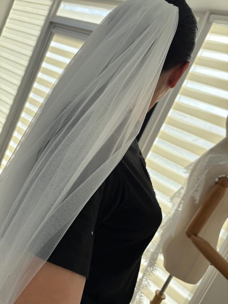 May include: A sheer white bridal veil, draped over the head and shoulders. The veil is made of a delicate, flowing fabric, creating a soft, ethereal look. The image is taken from a side angle, showcasing the veil's texture and design.