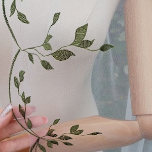 May include: White sheer fabric with green embroidered leaves. The fabric is draped over a wooden mannequin.