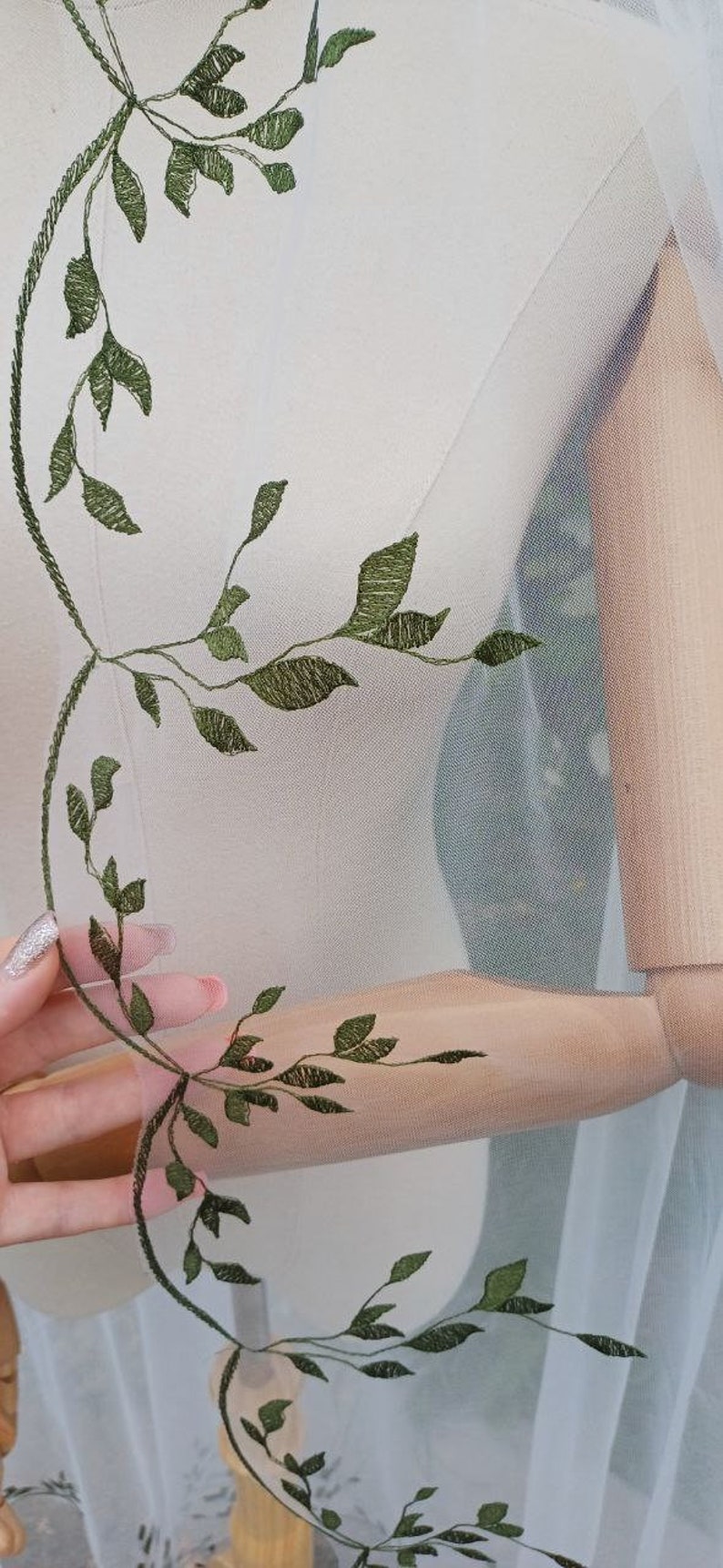 May include: White sheer fabric with green embroidered leaves. The fabric is draped over a wooden mannequin.