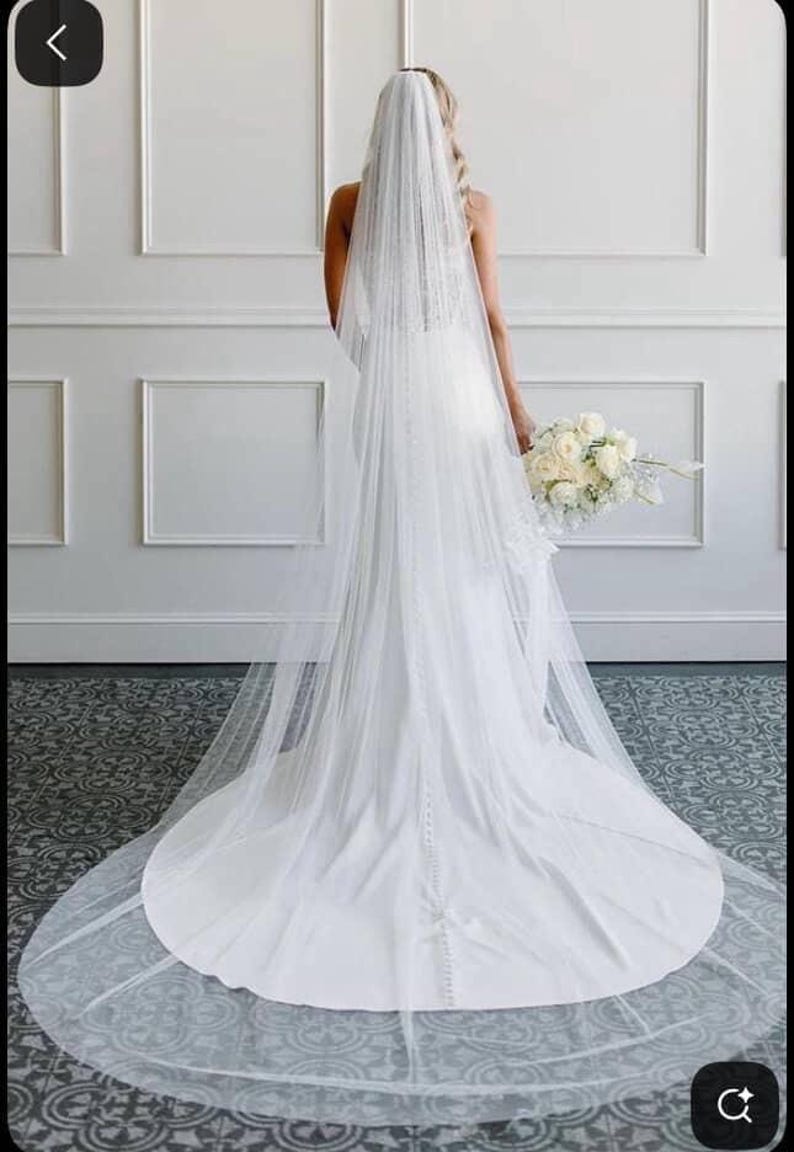 May include: A white wedding dress with a long train and a sheer veil. The bride is holding a bouquet of white roses. The dress has a fitted bodice and a flowing skirt. The veil is long and delicate.