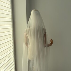 May include: A sheer, ivory-colored bridal veil cascading down a mannequin. The veil is long and flowing, creating a soft, ethereal look. The mannequin is positioned near a window with blinds, casting soft light on the veil.
