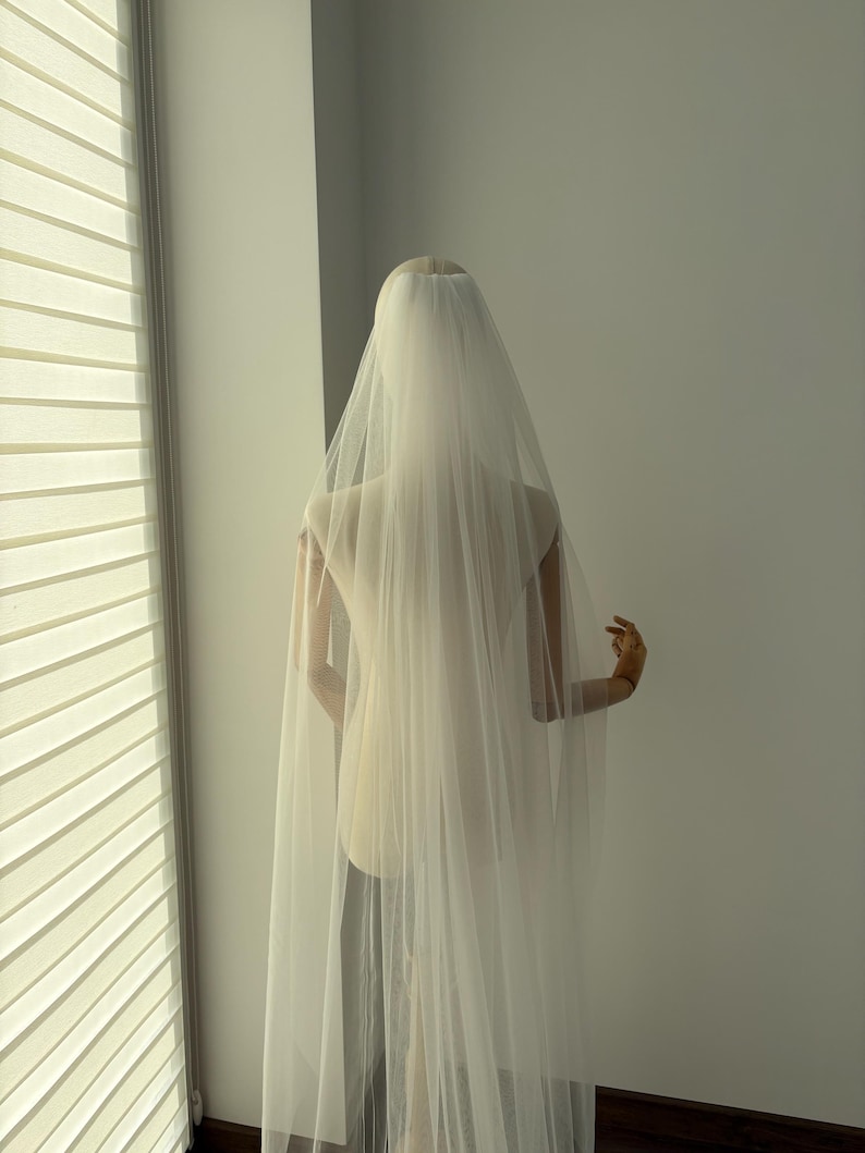 May include: A sheer, ivory-colored bridal veil cascading down a mannequin. The veil is long and flowing, creating a soft, ethereal look. The mannequin is positioned near a window with blinds, casting soft light on the veil.