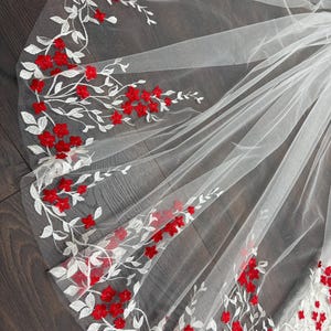 Könnte beinhalten: Ein weißer Brautschleier mit floralem Design. Der Schleier hat gestickte weiße Zweige mit roten Blüten. Der Schleier besteht aus einem transparenten, leichten Stoff. Das florale Design befindet sich an den Rändern des Schleiers.