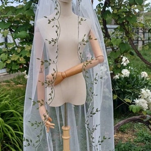 May include: A white tulle wedding veil with green embroidered leaves. The veil is draped over a wooden mannequin and is displayed against a backdrop of green foliage.