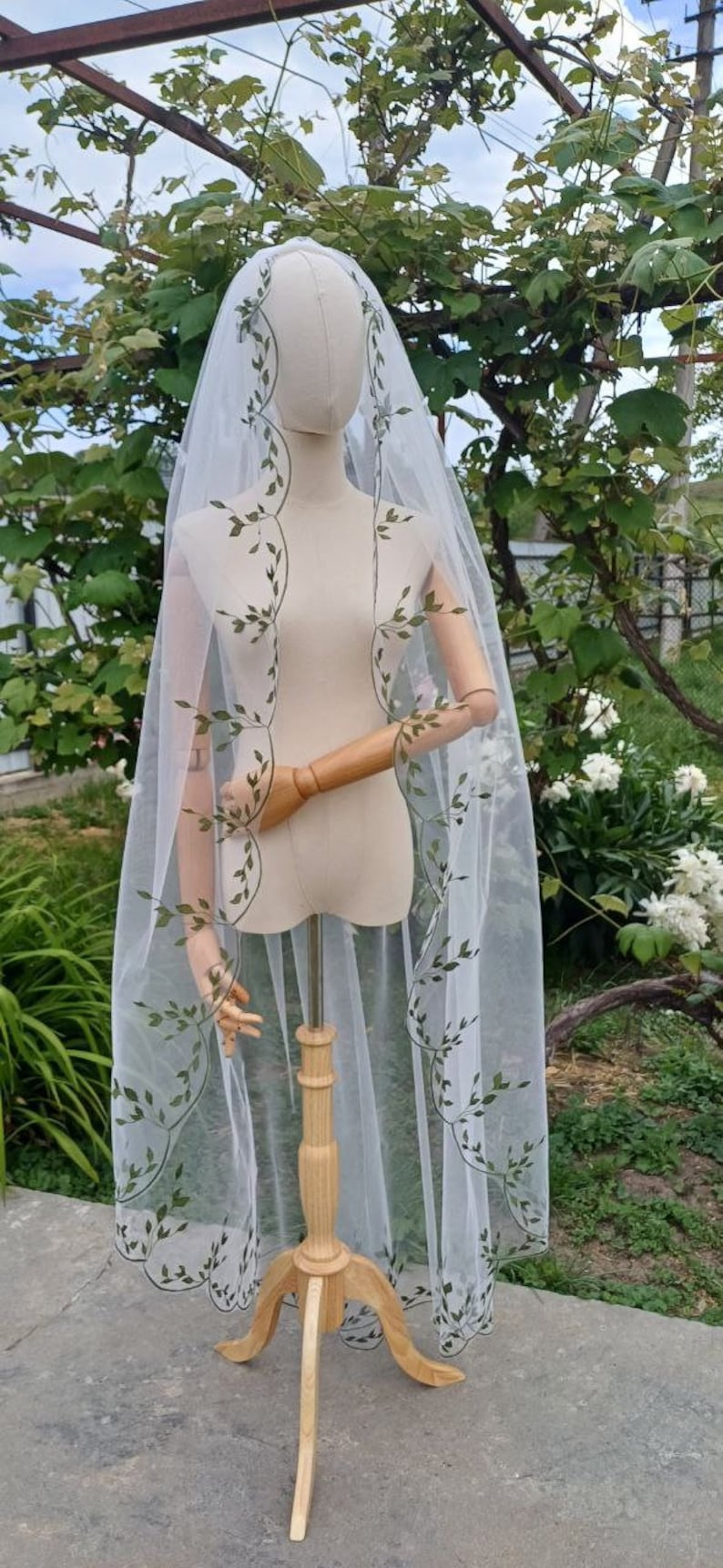 May include: A white tulle wedding veil with green embroidered leaves. The veil is draped over a wooden mannequin and is displayed against a backdrop of green foliage.