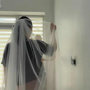 May include: A sheer white bridal veil is held up in front of a person wearing a gray t-shirt. The veil is long and flowing, with a delicate texture. The person is standing in front of a window with white blinds.