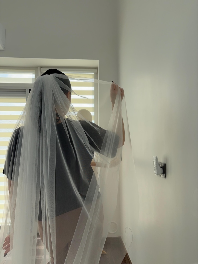May include: A sheer white bridal veil is held up in front of a person wearing a gray t-shirt. The veil is long and flowing, with a delicate texture. The person is standing in front of a window with white blinds.