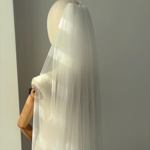 May include: A sheer, white bridal veil is draped over a mannequin head and shoulders. The veil is made of a lightweight, flowing fabric, and it cascades down the back. The mannequin is on a wooden stand, and the background is a plain wall.