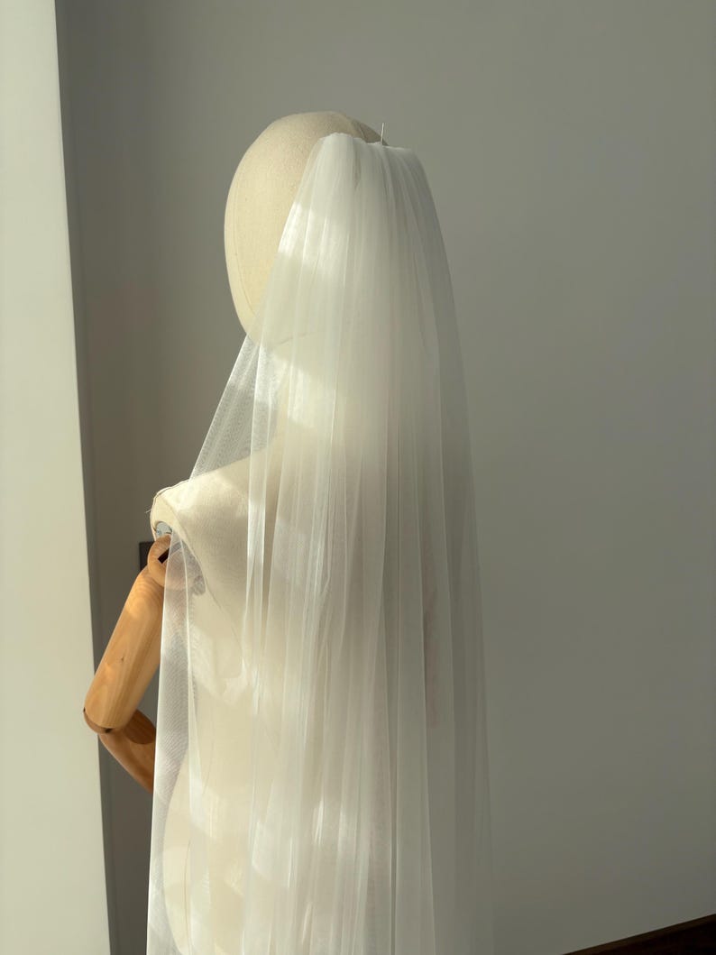 May include: A sheer, white bridal veil is draped over a mannequin head and shoulders. The veil is made of a lightweight, flowing fabric, and it cascades down the back. The mannequin is on a wooden stand, and the background is a plain wall.
