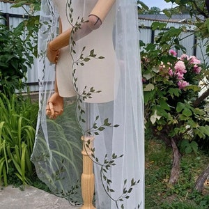 May include: White bridal veil with a delicate green floral pattern. The veil is draped over a wooden mannequin and has a scalloped edge.