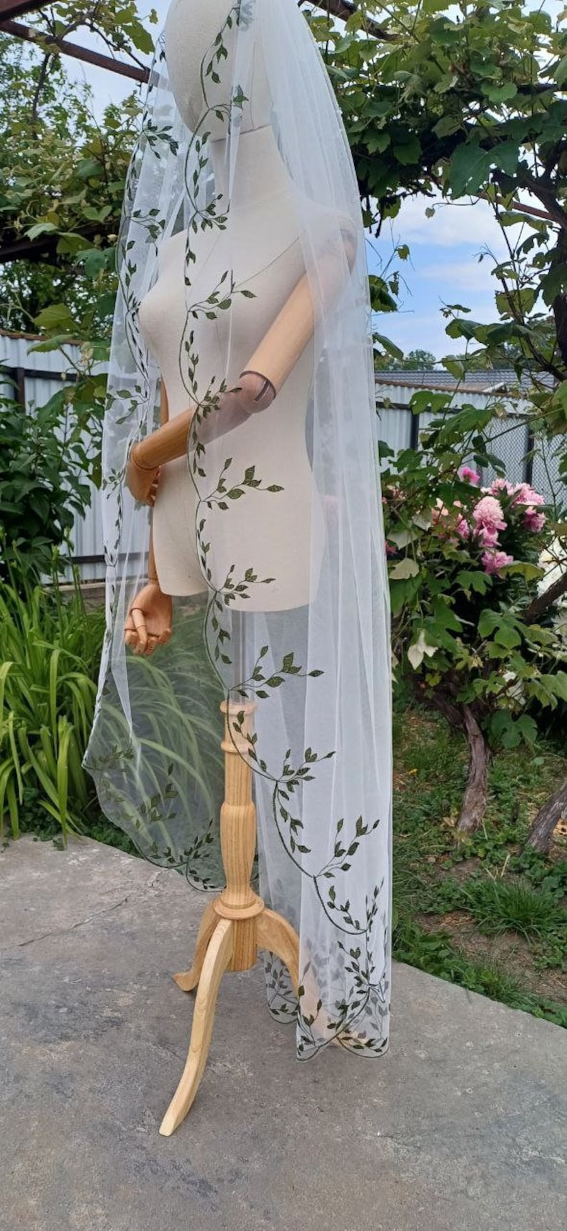 May include: White bridal veil with a delicate green floral pattern. The veil is draped over a wooden mannequin and has a scalloped edge.