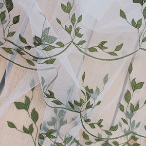 May include: White sheer fabric with a delicate green leaf embroidery pattern. The fabric is draped over a wooden surface.