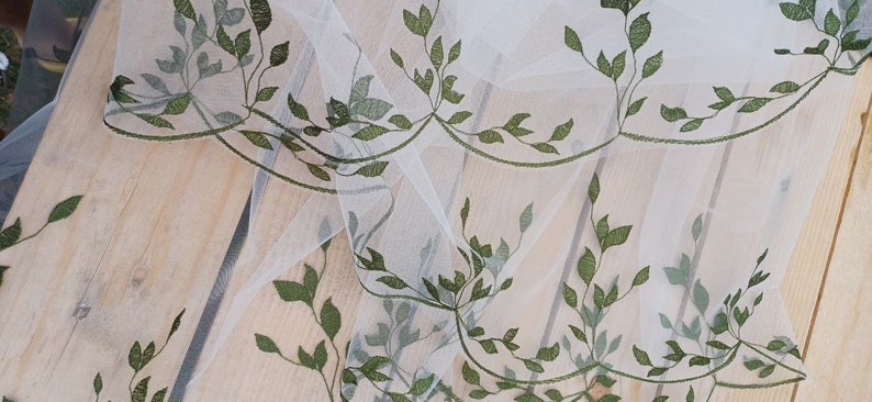 May include: White sheer fabric with a delicate green leaf embroidery pattern. The fabric is draped over a wooden surface.