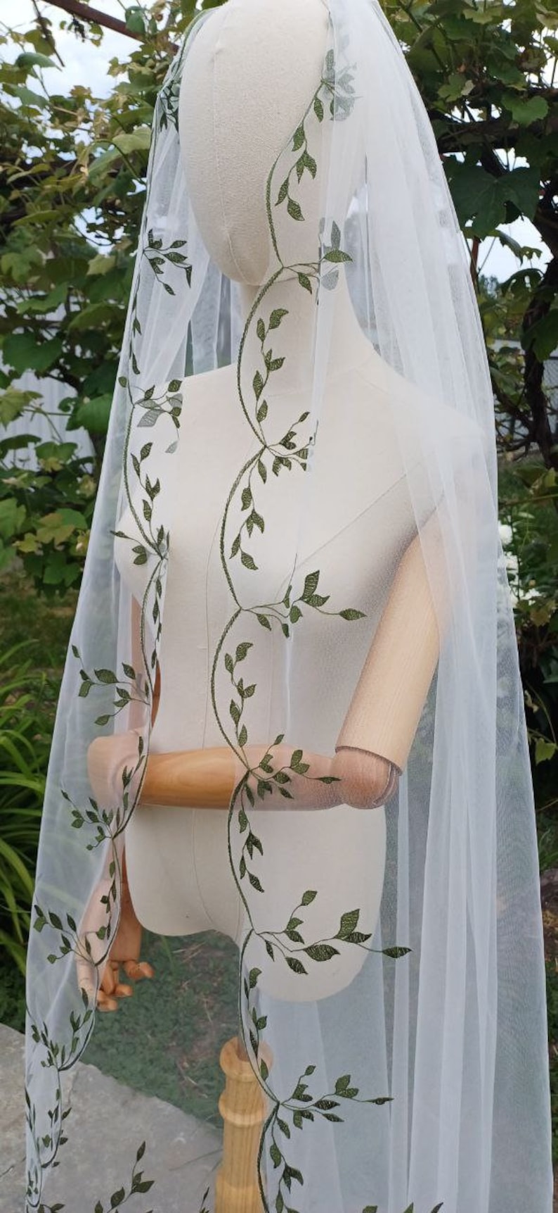 May include: White bridal veil featuring a delicate green leaf embroidery pattern. The veil is sheer and flowing, with a soft, romantic look.