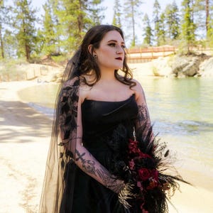 Könnte beinhalten: Schwarzes trägerloses Brautkleid mit passendem Schleier mit floralen Details. Die Braut hält einen Strauß aus dunkelroten Rosen und schwarzen Federn. Im Hintergrund sind Strand und Bäume zu sehen.