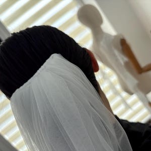 May include: A close-up shot of a white tulle veil, draped over black hair. The veil is gathered at the top and falls in soft folds. A mannequin is visible in the background, suggesting bridal or formal wear.