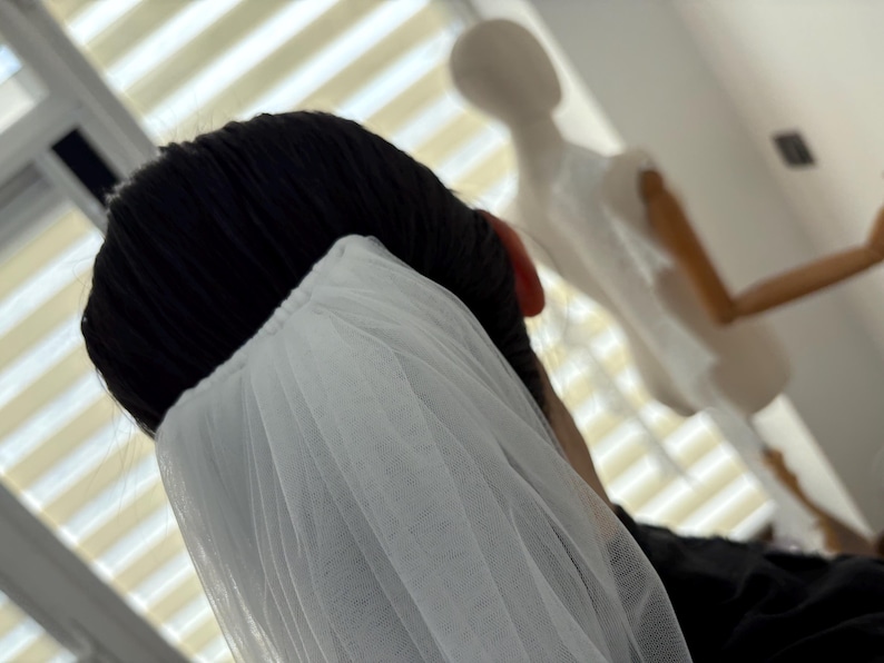 May include: A close-up shot of a white tulle veil, draped over black hair. The veil is gathered at the top and falls in soft folds. A mannequin is visible in the background, suggesting bridal or formal wear.