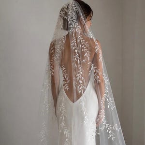 May include: A long, sheer white bridal veil featuring delicate, embroidered white leaf and vine patterns throughout the fabric. The veil is draped over a simple, sleeveless white gown with a deep, open back design, creating a layered appearance.